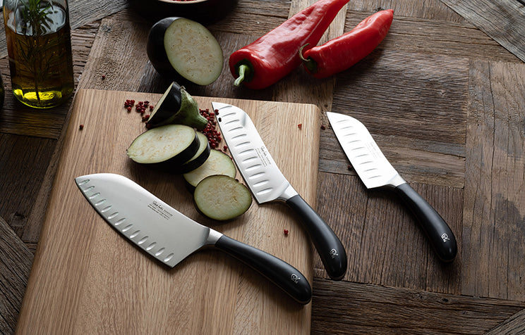 Three Signature Santoku Kitchen Knives on a wooden cutting board with sliced vegetables.
